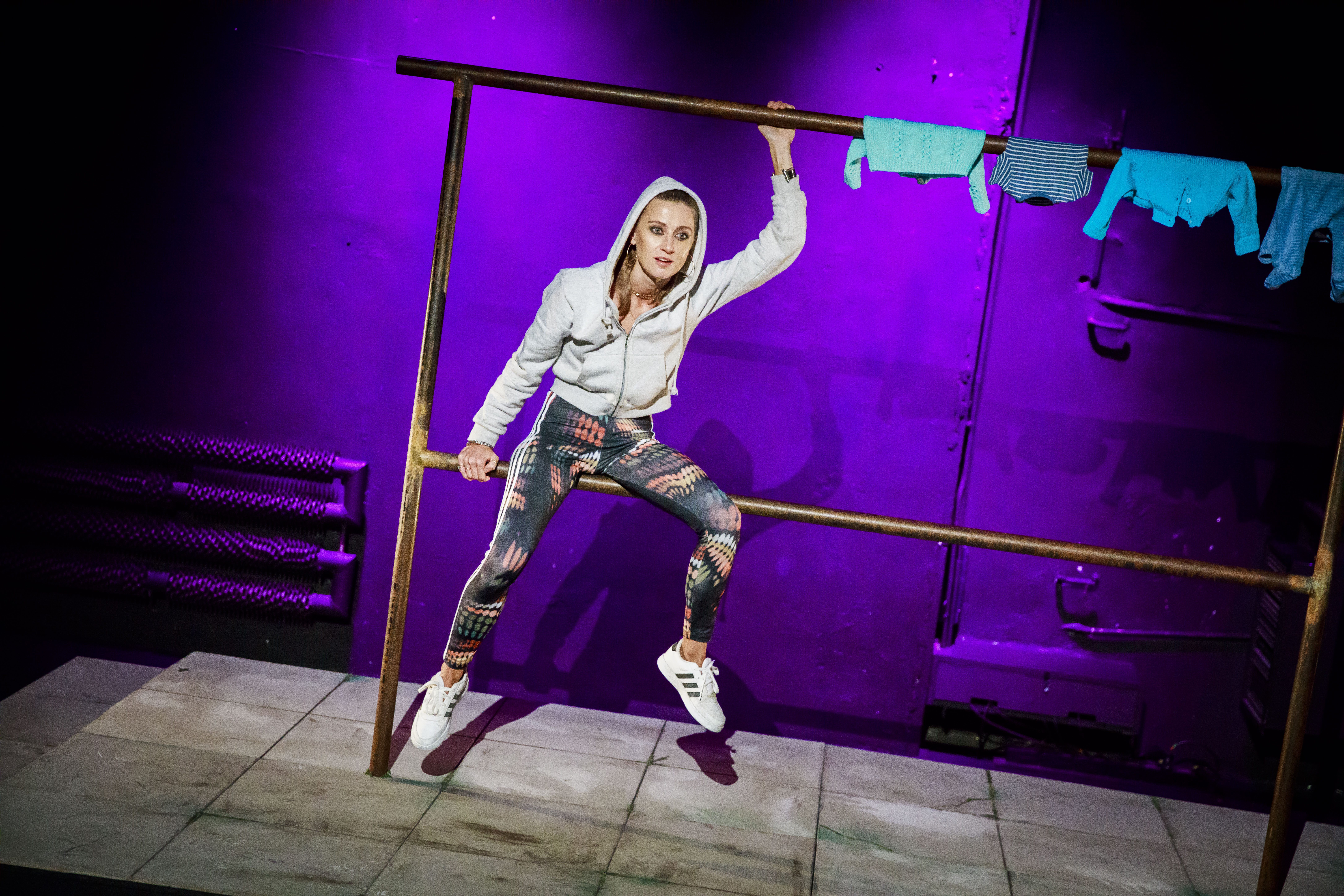 zdjęcie ze spektaklu „Ifigenia ze Splott". Aktorka Agnieszka Skrzypczak siedzi na trzepaku, na którym wiszą dziecięce ubrania. Ma na sobie szarą bluzę od dresu oraz opięte leginsy, na stopach adidasy. Na głowie ma kaptur. Jej oczy są mocno podkreślone makijażem. Ma kolczyki koła. Scena jest oświetlona fioletowym światłem.