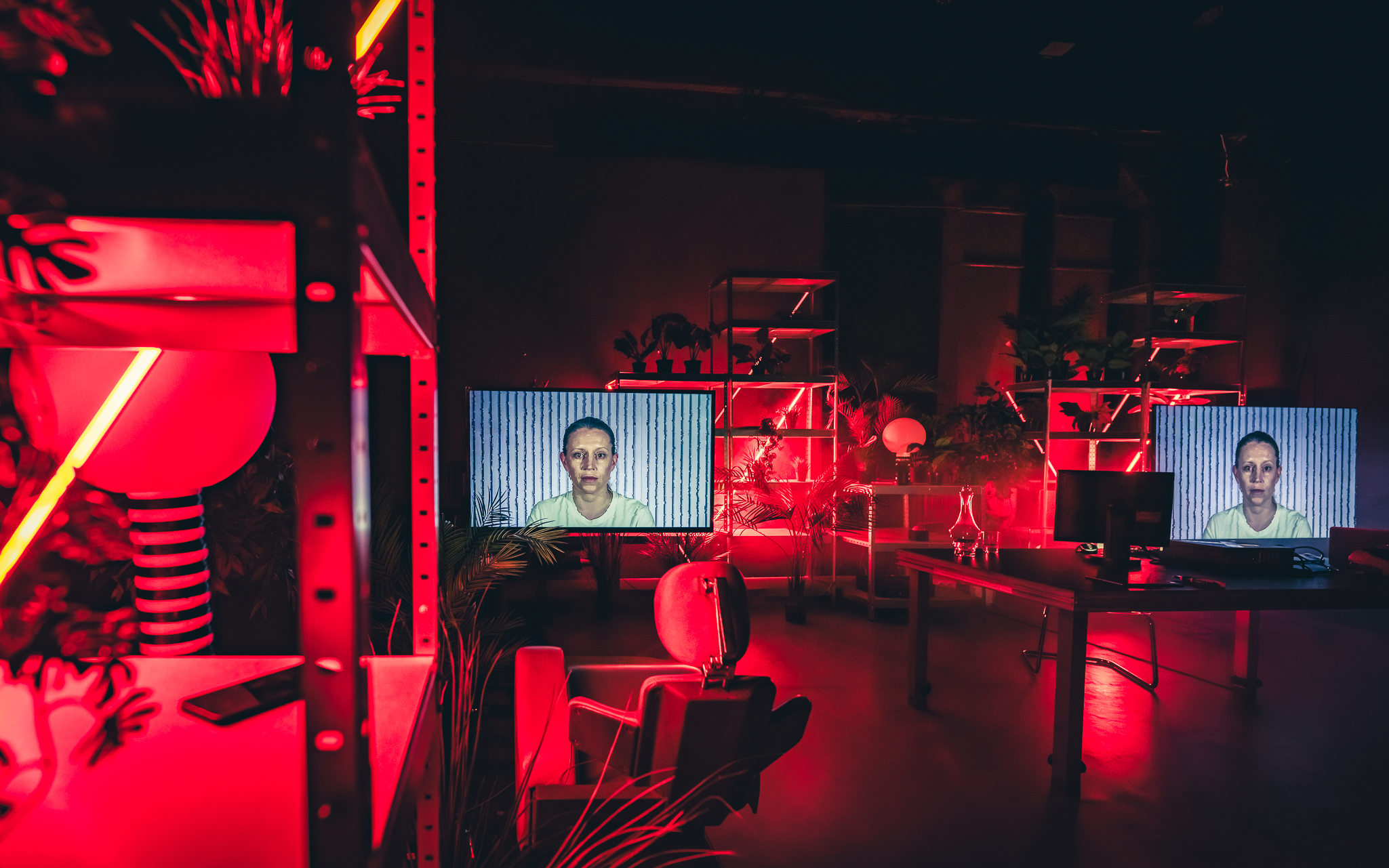 Mroczna, laboratoryjna przestrzeń sceniczna skąpana jest w intensywnym, czerwonym świetle nieonowym. Na dwóch dużych monitorach rozstawionych wśród regałów z roślinami wyświetlany jest ten sam portret kobiety.