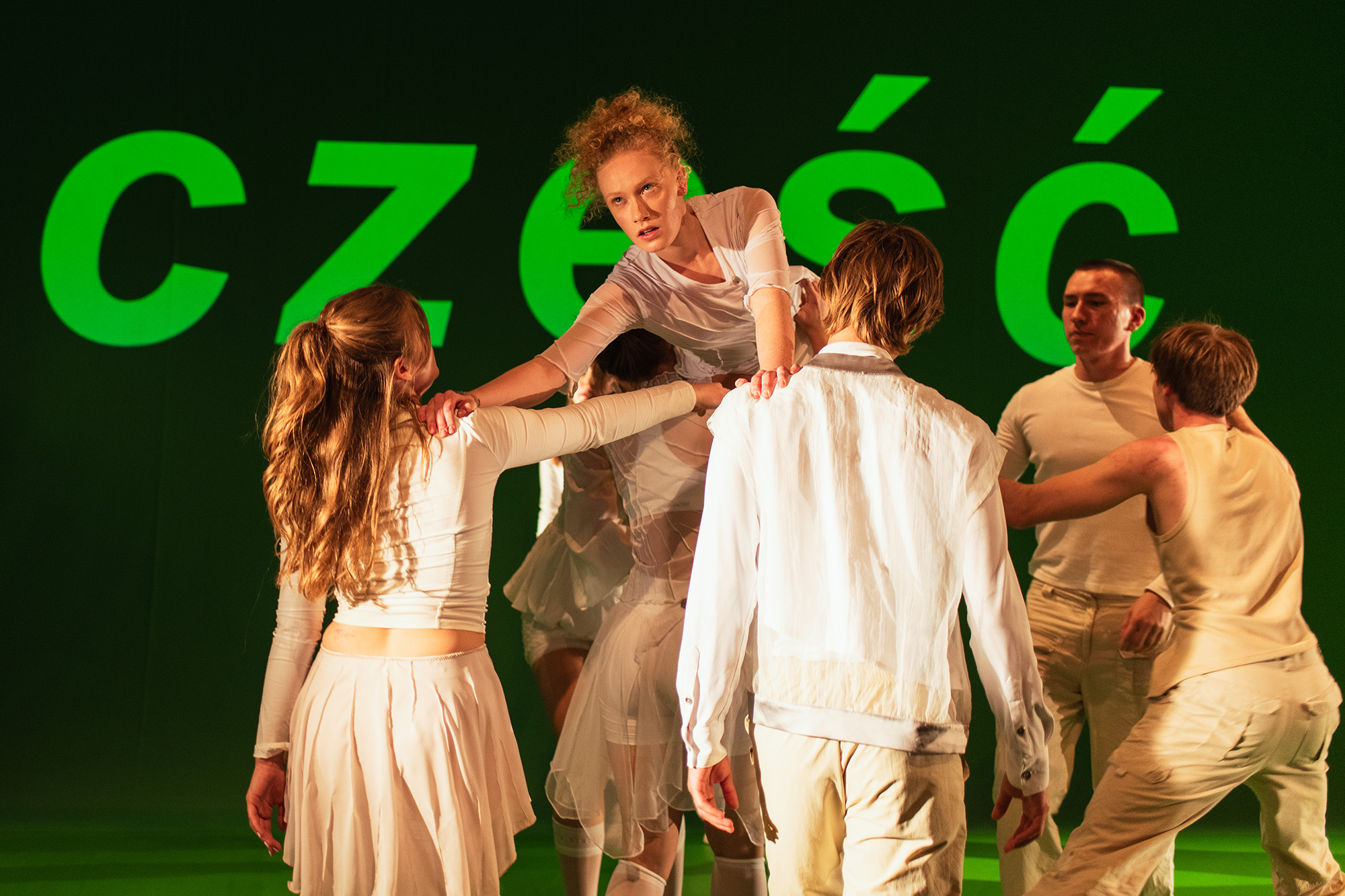 zdjęcie ze spektaklu „Stany podgorączkowe" z AST w Krakowie. Na zdjęciu widać siódemkę aktorów. Na pierwszym planie troje z nich (jednak kobieta i dwaj mężczyźni) stoją tyłem. Na nich i na barkach pozostałych wspina się jedna aktorka. Z tyłu, za aktorami, widać zielony napis "cześć"