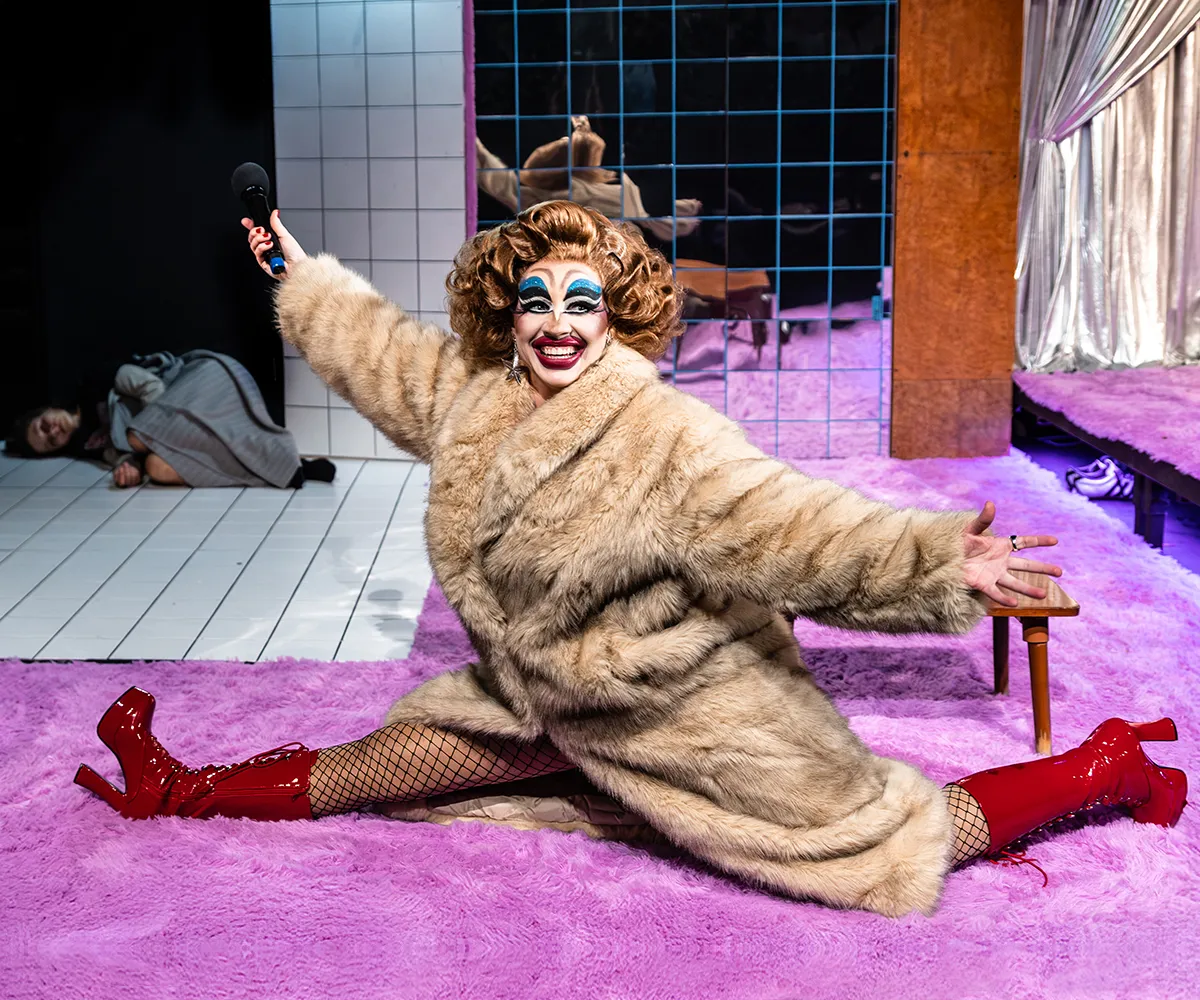 Zdjęcie ze spektaklu „Czas na wielki numer". Na fotorafii widać aktorkę Matyldę Paszczenko w charakteryzacji na drag queen. Ma na sobie gruby płaszcz. Robi szpagat. W tle pod drzwiami leży aktorka Natalia Klepacka. Autorką zdjęcią jest Natalia Kabanow