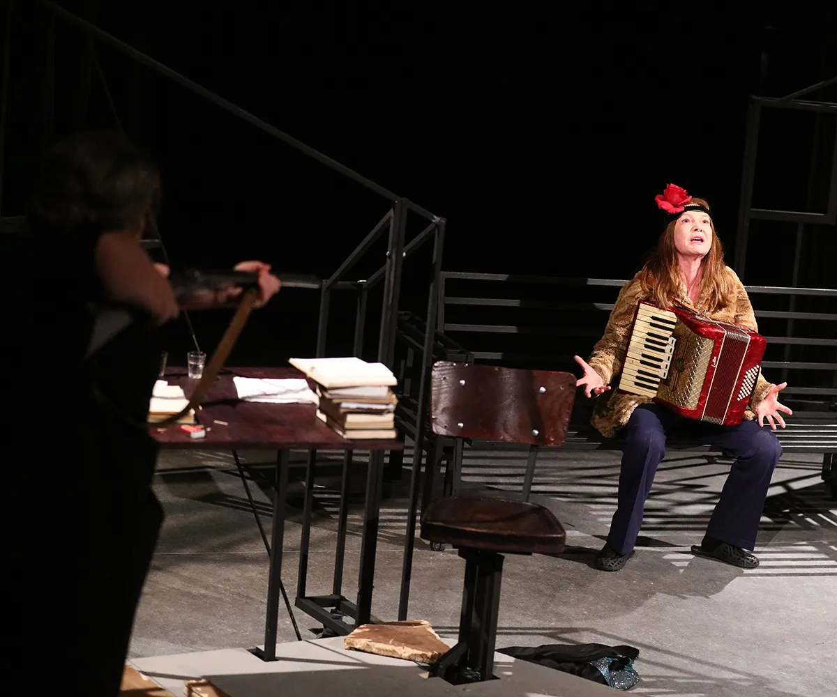 Scena teatralna przedstawiająca dramatyczny moment. Po prawej stronie siedzi kobieta z rozłożonymi rękami, grając na czerwono-złotej akordeonie. Ma na sobie ciemne spodnie, wzorzystą bluzkę lub narzutę oraz opaskę z czerwonym kwiatem we włosach. Z lewej strony, tyłem do widza, stoi druga postać celująca w nią z broni przypominającej strzelbę. Na pierwszym planie znajduje się ciemne drewniane biurko z leżącymi na nim dokumentami i książkami oraz krzesło obrotowe. W tle widoczna jest metalowa konstrukcja scen