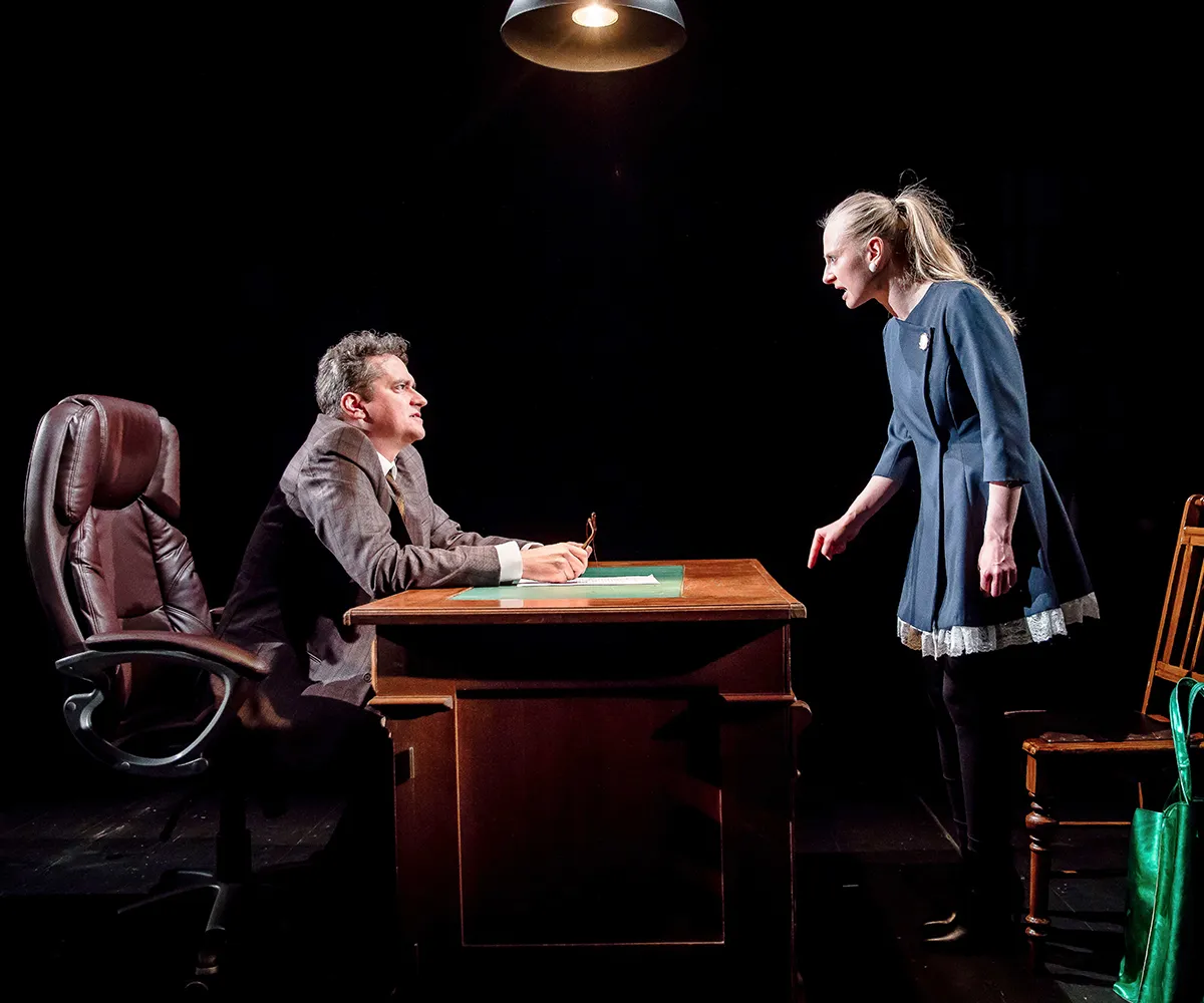 Scena teatralna w ciemnym oświetleniu, gdzie dwoje aktorów odgrywa konfrontację przy biurku. Po lewej stronie, siedząc w dużym brązowym skórzanym fotelu za drewnianym biurkiem, znajduje się mężczyzna z siwymi włosami, ubrany w tweedową marynarkę i koszulę. Jego dłonie oparte są o blat, a wzrok skierowany jest na kobietę. Stojąca po prawej stronie kobieta w niebieskiej sukience i z włosami spiętymi w koński ogon, jest pochylona i krzyczy, wskazując palcem na mężczyznę. Biurko jest zagracone, a w tle, po praw