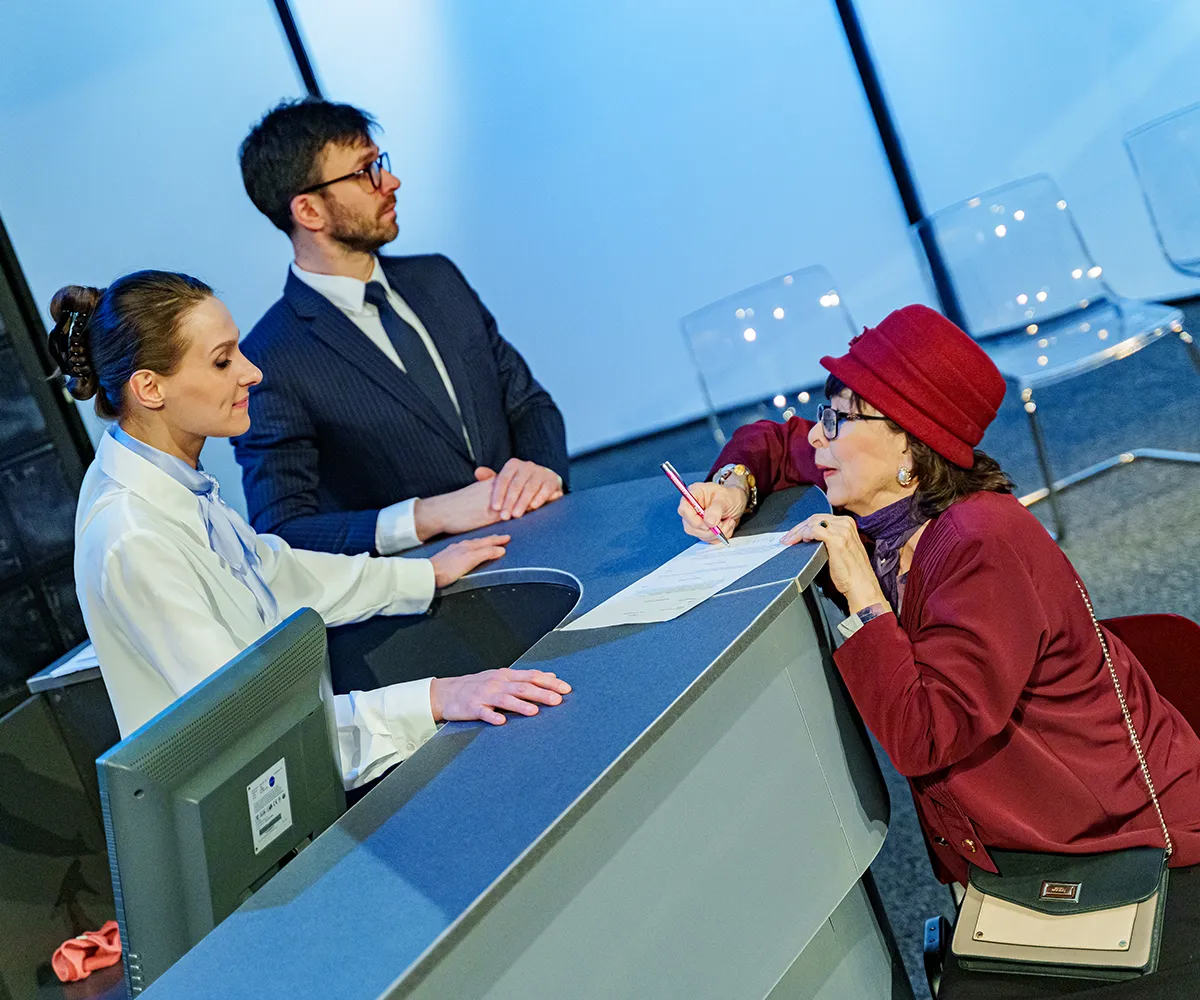 Pracowniczka w białej bluzce z apaszką obsługuje starszą kobietę w bordowym kapeluszu i żakiecie, która podpisuje dokument. Obok stoi mężczyzna w garniturze.