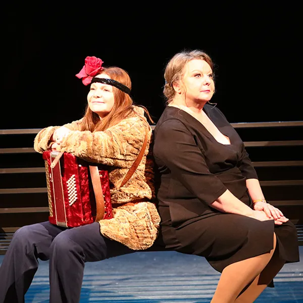Scena teatralna przedstawiająca dramatyczny moment. Po prawej stronie siedzi kobieta z rozłożonymi rękami, grając na czerwono-złotej akordeonie. Ma na sobie ciemne spodnie, wzorzystą bluzkę lub narzutę oraz opaskę z czerwonym kwiatem we włosach. Z lewej strony, tyłem do widza, stoi druga postać celująca w nią z broni przypominającej strzelbę. Na pierwszym planie znajduje się ciemne drewniane biurko z leżącymi na nim dokumentami i książkami oraz krzesło obrotowe. W tle widoczna jest metalowa konstrukcja scen