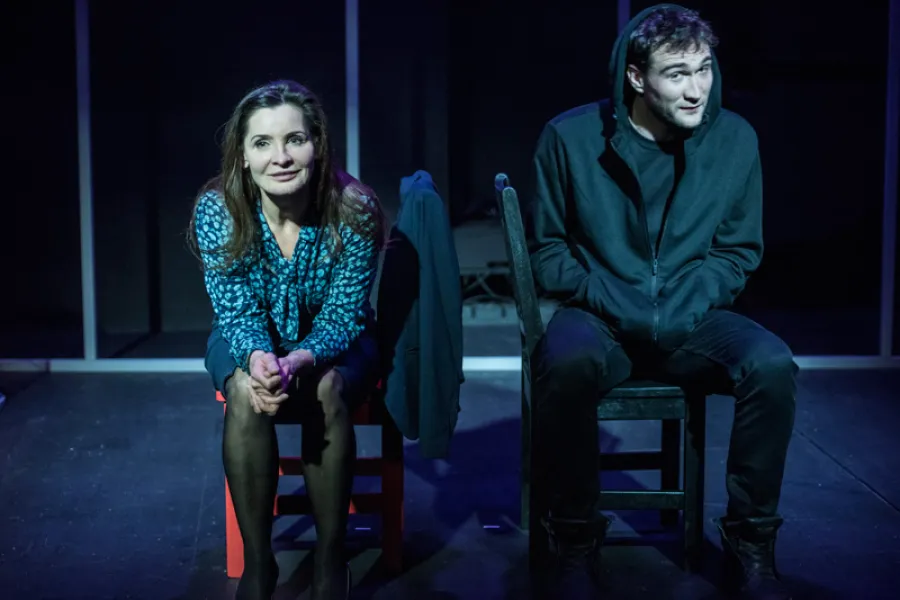 Kobieta w niebieskiej bluzce w kwiaty siedzi na czerwonym krześle, a obok niej na ciemnym krześle siedzi młody mężczyzna w czarnej bluze z kapturem. Obydwoje znajdują się na scenie, patrząc w stronę publiczności.