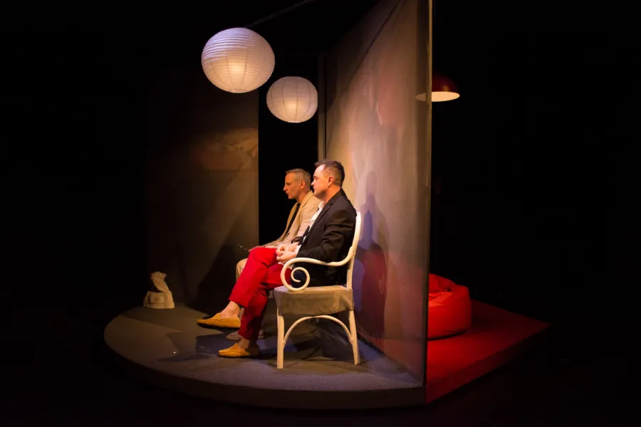 Dwóch mężczyzn siedzi na krzesłach na scenie pod wiszącymi kulistymi lampami. Sceneria ma teatralny charakter z ciemnymi ścianami i podestem.