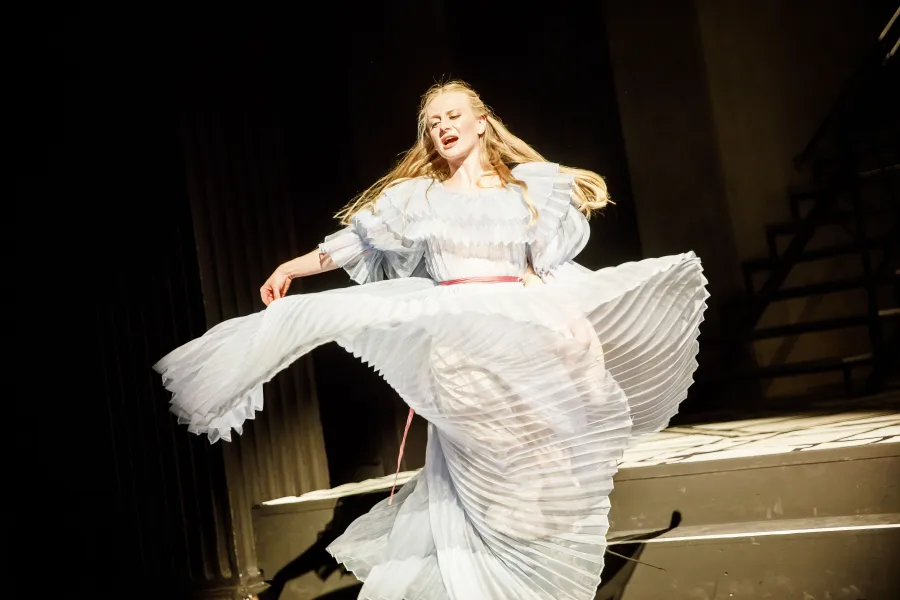 Blondynka w jasnej, zwiewnej sukni z plisowanym dołem wykonuje obrót w tańcu na scenie teatralnej. Wyraz jej twarzy i rozwiana spódnica nadają postaci niezwykłej dynamiki.