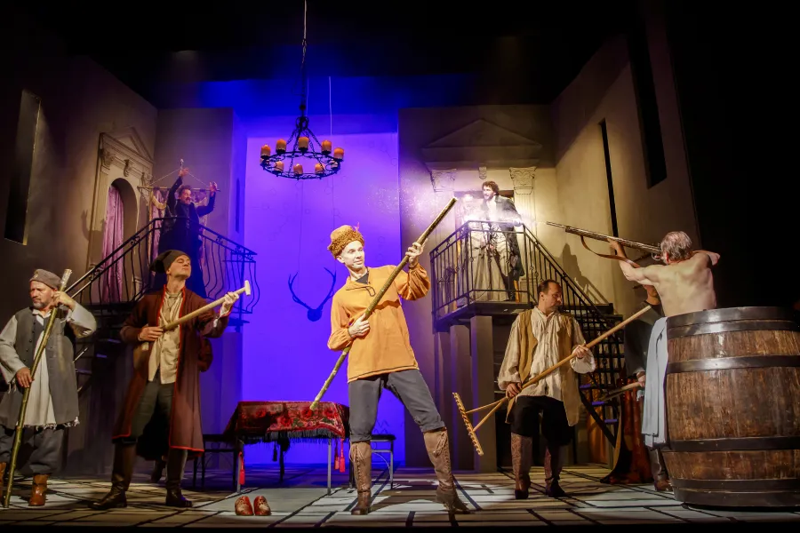 Dynamiczna scena teatralna przedstawia mężczyzn w dawnych strojach wiejskich i szlacheckich, trzymających w rękach broń palną, kije oraz grabie. Na pierwszym planie wyróżnia się mężczyzna w żółtej koszuli celujący z długiej strzelby, podczas gdy tło oświetlone jest intensywnym, niebieskim światłem.