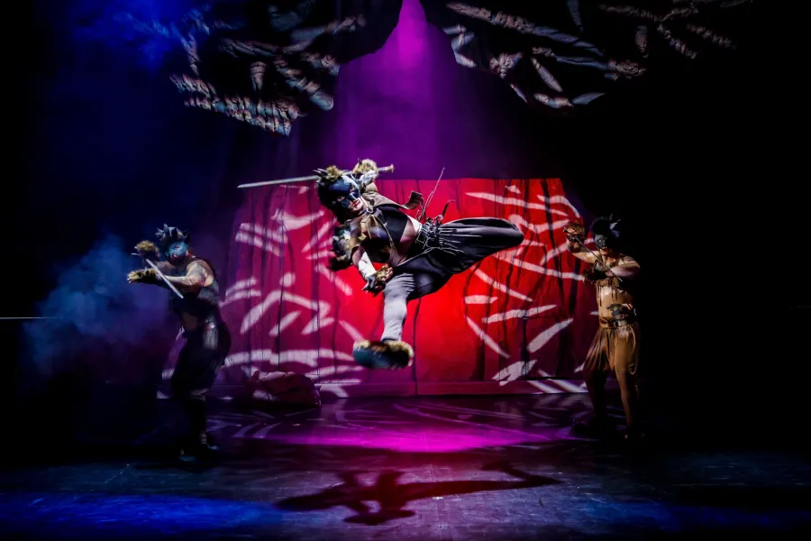 Troje wykonawców w kostiumach przypominających dzikie zwierzęta na scenie teatralnej; centralna postać w masce wykonuje dynamiczny skok w powietrzu z mieczem, podczas gdy dwaj pozostali stoją po bokach w pozach bojowych. Scena jest dramatycznie oświetlona niebieskim i fioletowym światłem punktowym, ze zwierzęcym motywem na tylnej kurtynie.