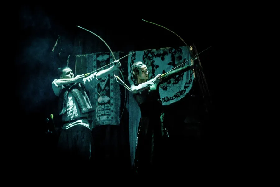 Dwie postacie, elfia kobieta i mężczyzna w stroju ludowym, celują z łuków na ciemnej scenie teatralnej przed zdobionymi tkaninami. Całość jest oświetlona chłodnym, niebiesko-zielonym światłem punktowym.