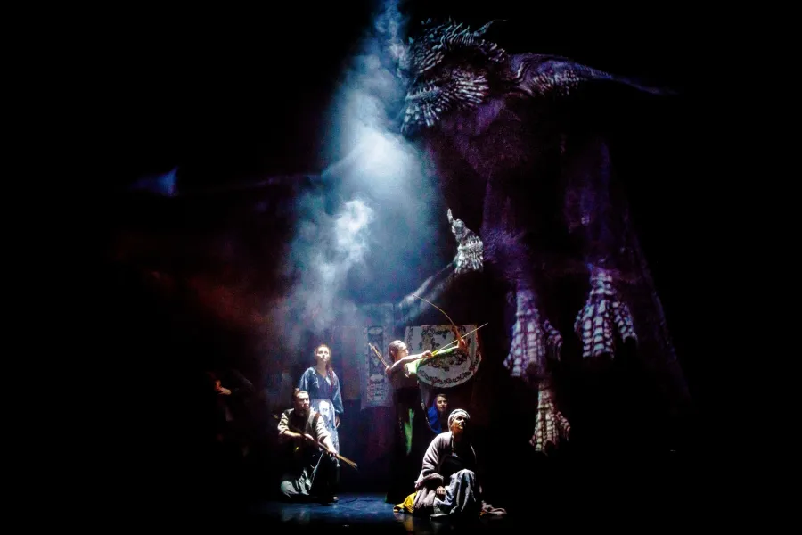 Ogromny, ciemny smok wznosi się nad grupą aktorów na spowitej dymem scenie teatralnej. Postaci na dole, w tym łuczniczka celująca w potwora, obserwują go z przerażeniem w strumieniu jasnego światła.