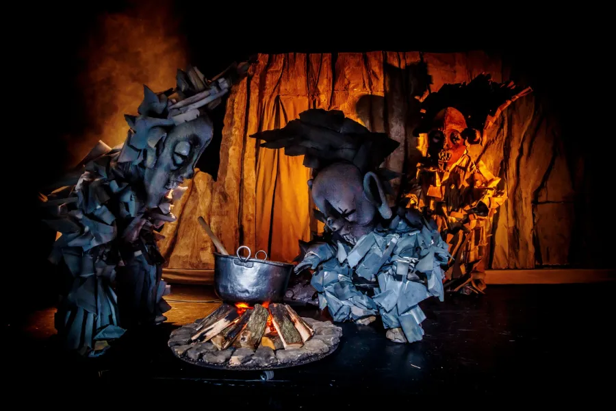 Trzy postaci w fantazyjnych, szarych i kamiennych kostiumach z dużymi maskami gotują w kociołku nad płonącym ogniskiem. Scena teatralna oświetlona jest ciepłym, pomarańczowym blaskiem ognia na ciemnym tle.
