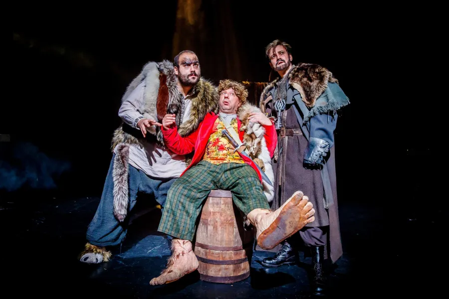 Trzech aktorów na mrocznej scenie teatralnej, jeden siedzi na beczce w barwnym stroju i z owłosionymi, nienaturalnie dużymi stopami. Obok niego stoją dwaj mężczyźni w kostiumach z futrem, jeden o zwierzęcych cechach twarzy, a drugi z brodą, wszyscy spoglądają z napięciem w górę.