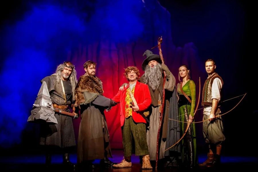 Grupa sześciu postaci w kostiumach nawiązujących do fantasy stoi na scenie teatralnej oświetlonej niebieskim i czerwonym światłem. W centrum uśmiechnięty mężczyzna w czerwonej marynarce i z owłosionymi stopami podaje rękę jednemu z towarzyszy, otoczony m.in. przez czarodzieja z laską i łuczniczkę.