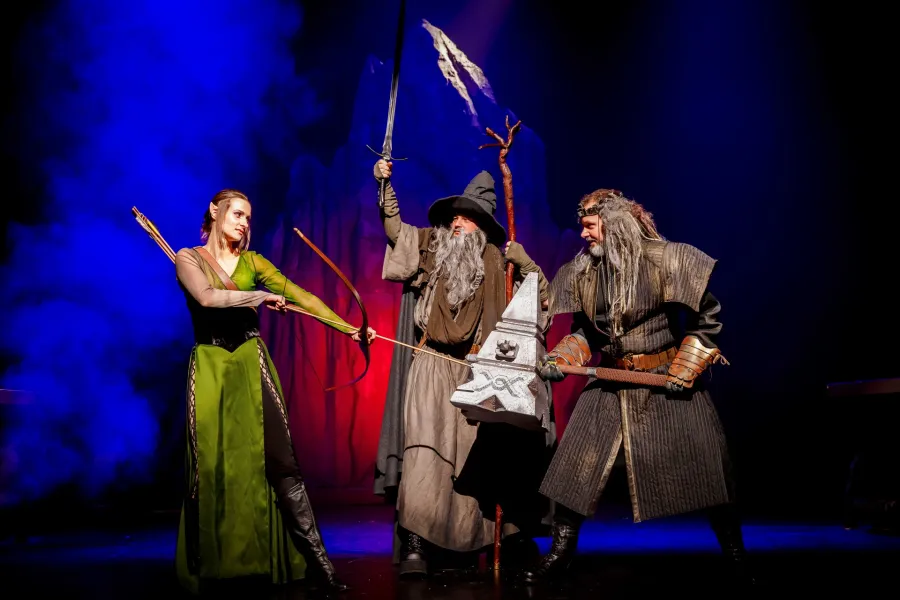 Troje aktorów w kostiumach fantasy – łuczniczka w zieleni, czarodziej z uniesionym mieczem i krasnoludzki król z wielkim młotem – pozuje w bojowych układach na scenie. Całość tła spowita jest dymem oraz intensywnym, niebiesko-czerwonym oświetleniem scenicznym.