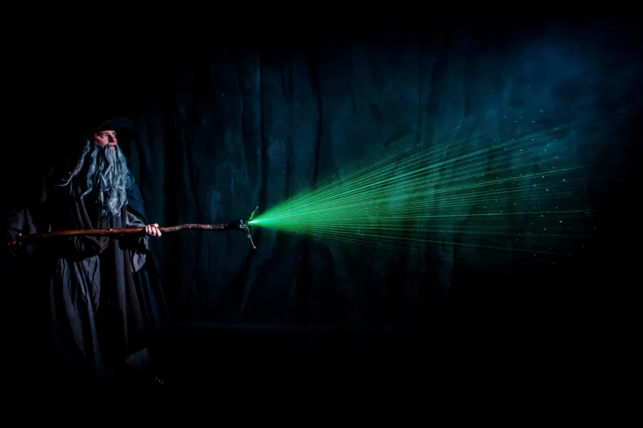 Aktor w kostiumie czarodzieja z długą, szarą brodą trzyma poziomą laskę, z której czubka wybiega intensywna wiązka zielonych promieni laserowych. Całość rozgrywa się na mocno zaciemnionej scenie z ciemnym, skalistym tłem.