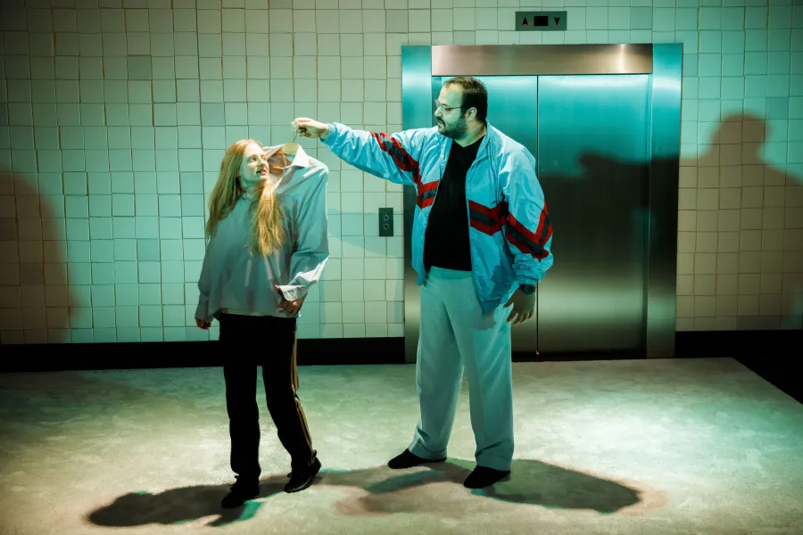 Na scenie teatralnej mężczyzna w jasnoniebieskim dresie trzyma na wieszaku koszulę, którą nakłada na stojącą obok kobietę z jasnymi włosami. W tle widoczna jest wykafelkowana ściana oraz drzwi windy.