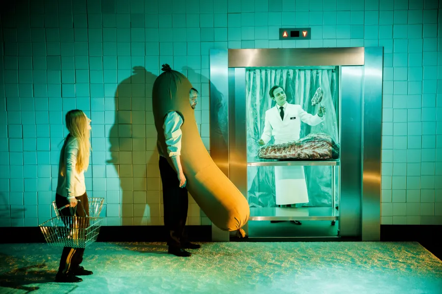 Na scenie teatralnej kobieta z koszykiem zakupowym patrzy na mężczyznę przebranego za gigantyczną parówkę. W tle, w otwieranym jak winda boksie z foliową kurtyną, stoi postać w białym fartuchu rzeźnika, która trzyma mięso.