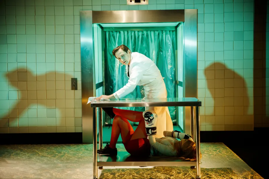 Na scenie teatralnej mężczyzna w białym fartuchu przeciera metalowy stół, pod którym leży kobieta w czerwonych rajstopach i rękawicach bokserskich. W tle widoczna jest kabina z zieloną kurtyną foliową oraz wykafelkowana ściana.