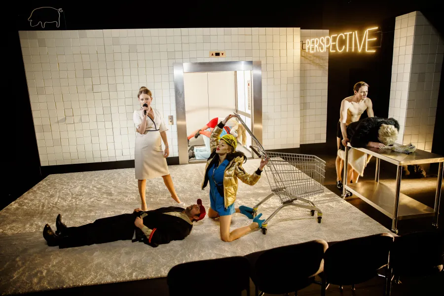 Na scenie teatralnej grupa aktorów odgrywa dynamiczną scenę na tle kafelkowej ściany z napisem "PERSPECTIVE" oraz windą. Wokół leżącego na podłodze mężczyzny i klęczącej kobiety z wózkiem sklekowym znajdują się inne postacie, w tym śpiewająca kobieta z mikrofonem.