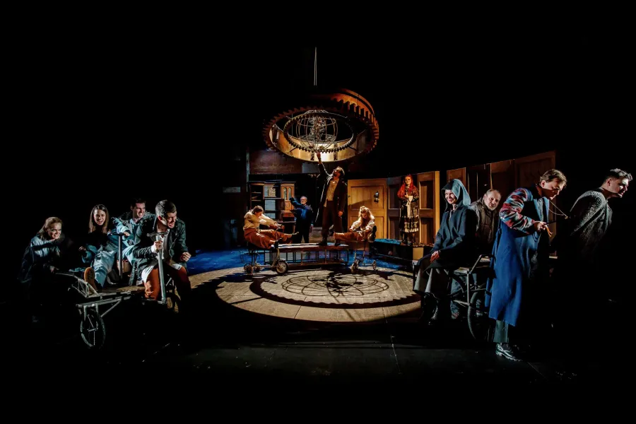 Grupa aktorów w zróżnicowanych kostiumach jest rozmieszczona na ciemnej scenie teatralnej wokół oświetlonego koła. Nad centralną częścią wznosi się duży, metalowy żyrandol w kształcie koła zębatego ze sferycznym elementem wewnątrz.