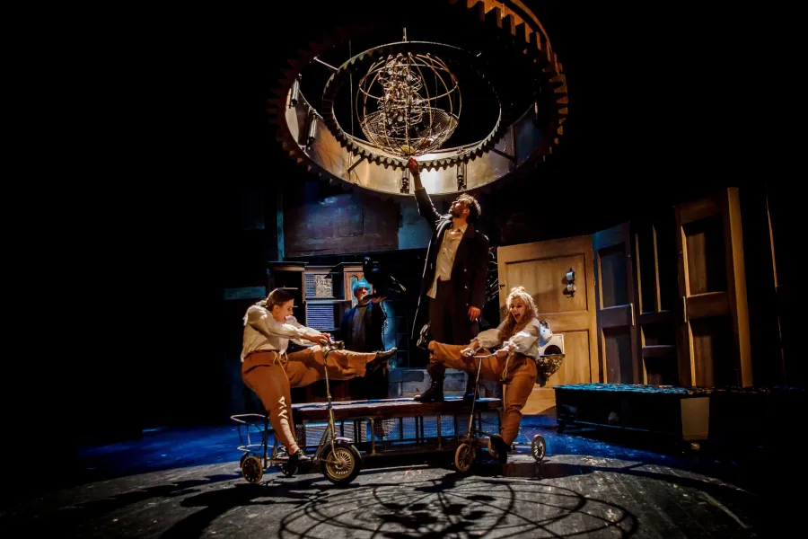 Cztery osoby występują na mrocznej scenie teatralnej ze steampunkową dekoracją, na której widać duże, metalowe koło zębate z sferyczną konstrukcją wewnątrz, wiszące nad nimi. Dwie performerki na pierwszym planie jadą na zabytkowych trójkołowcach, a mężczyzna w centrum, stojąc na platformie, sięga ręką w stronę wiszącej konstrukcji.