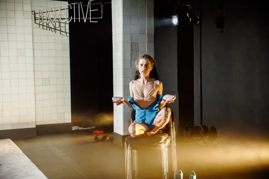 Na scenie teatralnej kobieta w bieliźnie i rozpiętym niebieskim stroju siedzi w kucki na stosie krzeseł, trzymając w dłoniach jasne przedmioty przypominające implanty piersi. W tle widoczny jest napis "PERSPECTIVE" oraz kafelkowana ściana.