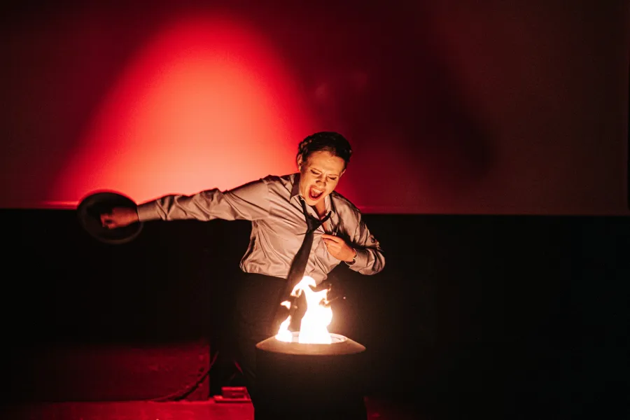 Kobieta w białej koszuli i krawacie krzyczy ekspresyjnie, stojąc nad płonącym pojemnikiem na ciemnej scenie teatralnej. W tle widać duży ekran oświetlony jasnym, czerwonym światłem, a kobieta trzyma w jednej ręce uniesiony talerz.