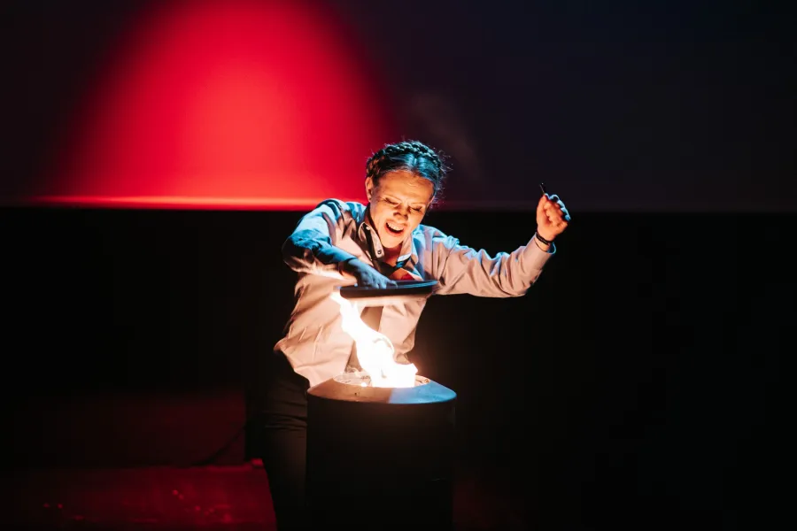 Kobieta w białej koszuli i krawacie krzyczy emocjonalnie, stojąc nad płonącym pojemnikiem na ciemnej scenie teatralnej. W tle widać duży ekran oświetlony jasnoczerwonym światłem, a w drugiej ręce kobieta trzyma nóż.