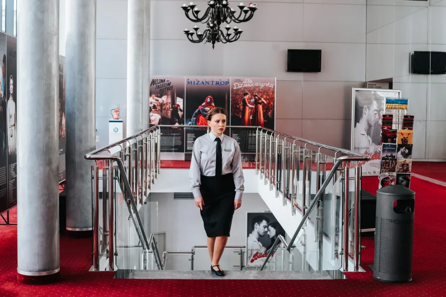 Kobieta w eleganckiej szarej koszuli, czarnym krawacie i ciemnej spódnicy schodzi po schodach w jasnym holu teatru. W tle widoczne są plakaty spektakli, czerwony dywan oraz ozdobny żyrandol pod sufitem.