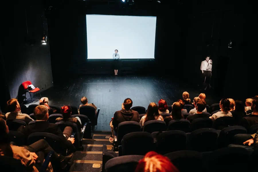 Widok z perspektywy widowni na kameralną, ciemną salę teatralną ze zgromadzoną publicznością. Na scenie, przed dużym białym ekranem, stoi samotna aktorka w białej koszuli i krawacie.