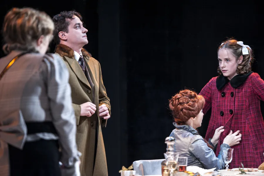 Na scenie teatralnej stoją cztery postacie, w tym stojący w centrum mężczyzna w płaszczu z futrzanym kołnierzem oraz dziewczynka w czerwonym płaszczu z białymi kokardami we włosach. Obok nich siedzą przy stole dwie inne kobiety, a cała scena rozgrywa się na ciemnym tle.