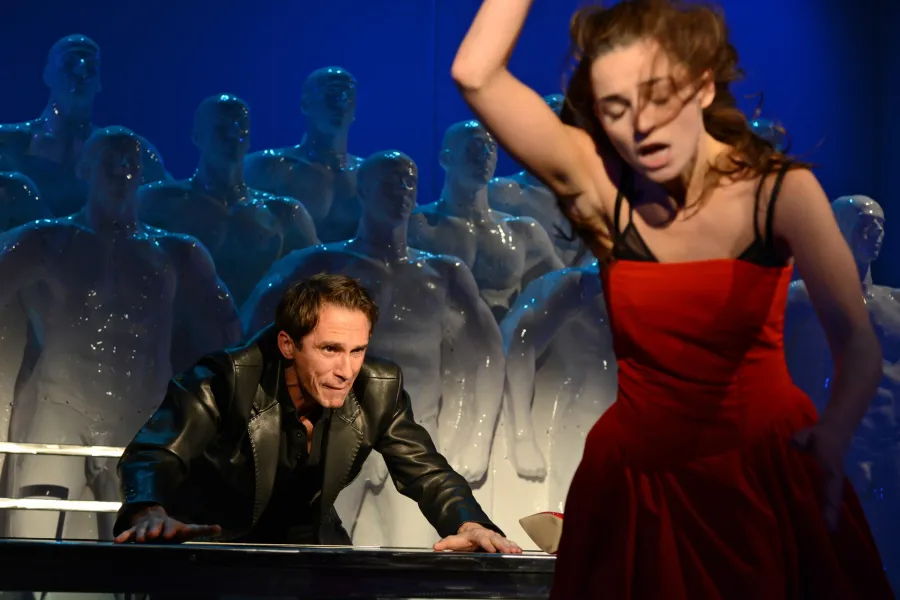 Na scenie teatralnej kobieta w czerwonej sukience i z rozpuszczonym włosami gwałtownie porusza się na pierwszym planie, jakby za chwilę miała upaść. W tle pochylony mężczyzna w skórzanej kurtce opiera się o stół, wpatrując się w kobietę na tle grupy białych manekinów.