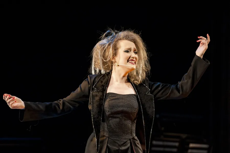 Kobieta z bujnymi włosami, ubrana w czarny gorset i żakiet, występuje na ciemnej scenie teatralnej z szeroko rozłożonymi rękami. Jej ekspresyjna twarz wyraża silne emocje podczas dramatycznego występu.