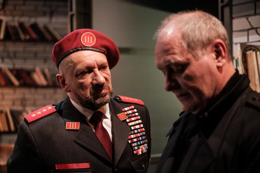 Mężczyzna w wojskowym mundurze i bordowym berecie z baretkami rozmawia z mężczyzną w czarnym ubraniu. W tle widać regał z książkami.