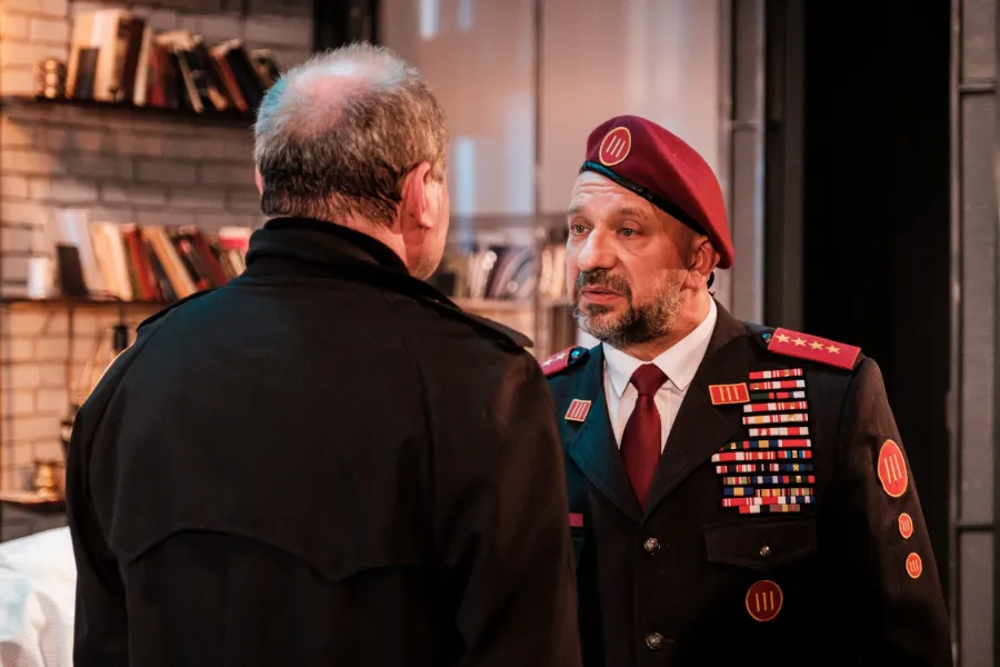 Mężczyzna w ciemnym płaszczu stoi tyłem, rozmawiając z wojskowym w mundurze i bordowym berecie, który ozdobiony jest licznymi baretkami i gwiazdkami. W tle widoczna jest jasna ściana z półkami wypełnionymi książkami.