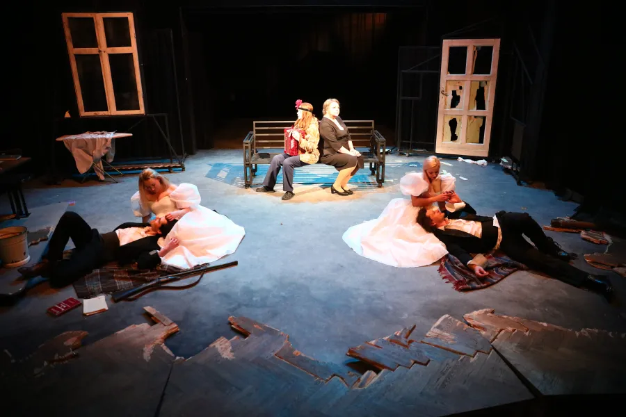 Na scenie teatralnej znajdują się dwie pary postaci w jasnych, ślubnych strojach, siedzące lub leżące na podłodze po bokach, oraz dwie kobiety siedzące tyłem do siebie na ławce w tle. Wokół widoczne są elementy dekoracji, w tym okna, drzwi i zniszczona podłoga.
