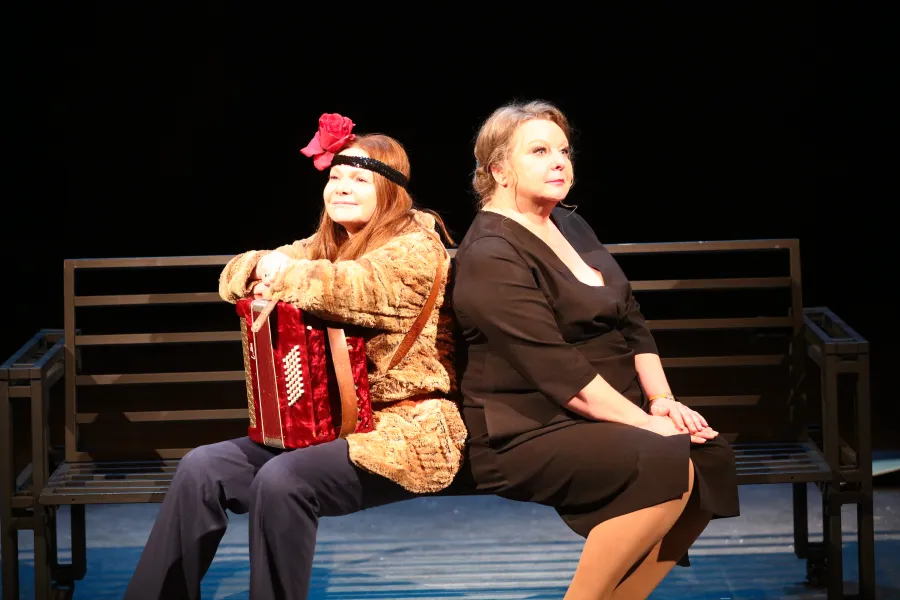 Dwie kobiety siedzą tyłem do siebie na ciemnej ławce na scenie teatralnej. Kobieta po lewej stronie ma na sobie futrzane okrycie wierzchnie i czerwoną różę we włosach oraz trzyma mały czerwony akordeon, a kobieta po prawej jest ubrana w elegancką czarną sukienkę.