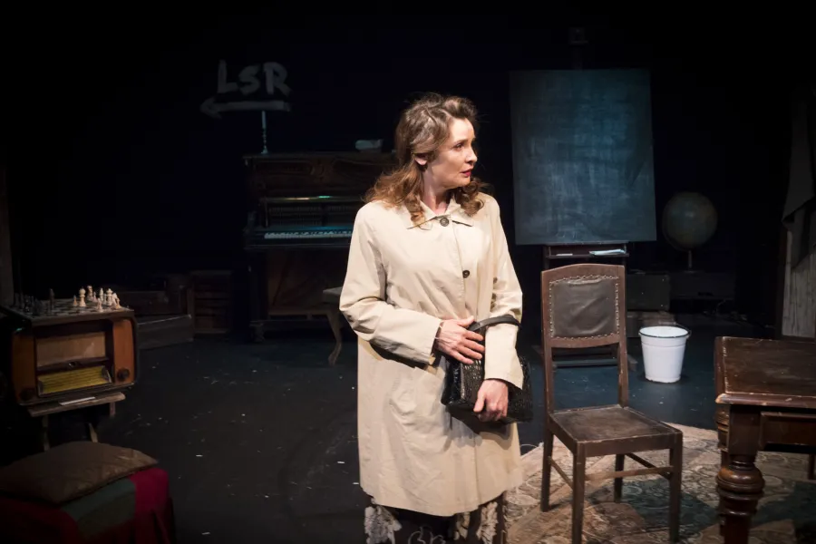 Kobieta w jasnym płaszczu trzyma torebkę i stoi na scenie teatralnej, rozglądając się z zatroskanym wyrazem twarzy. W tle widoczne są elementy scenografii, w tym pianino, krzesła, stolik oraz tablica.