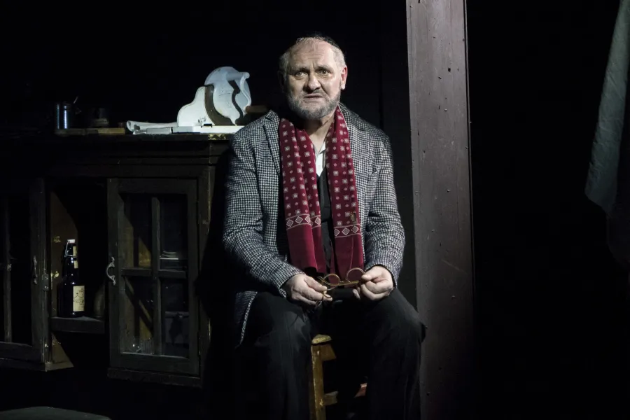 Mężczyzna w marynarce w kratę, czerwonym szaliku i jarmużce siedzi na krześle, trzymając w dłoniach okulary. W ciemnym tle po lewej stronie widoczna jest drewniana szafka z butelką.