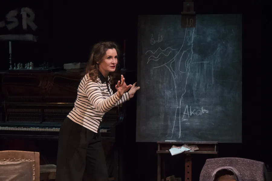 Kobieta w bluzce w paski stoi na scenie teatralnej i gestykuluje przed tablicą z narysowaną mapą. W tle widoczne jest pianino i inne rekwizyty.