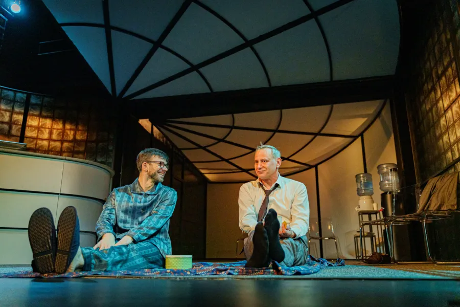 Na scenie teatralnej dwaj mężczyźni siedzą na podłodze rozłożonego koca w nowoczesnym biurowym wnętrzu. Jeden z nich ma na sobie piżamę w kratę, a drugi białą koszulę z krawatem i podwinięte nogawki spodni.