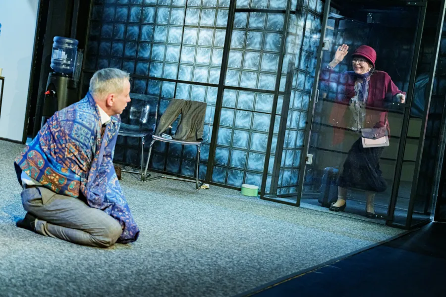 Na scenie teatralnej klęczący mężczyzna w ozdobnej narzutce patrzy w stronę stojącej za szklaną ścianą kobiety w czerwonym kapeluszu, która macha na pożegnanie. W tle widoczna jest ściana z luksferów oraz przezroczyste krzesła.