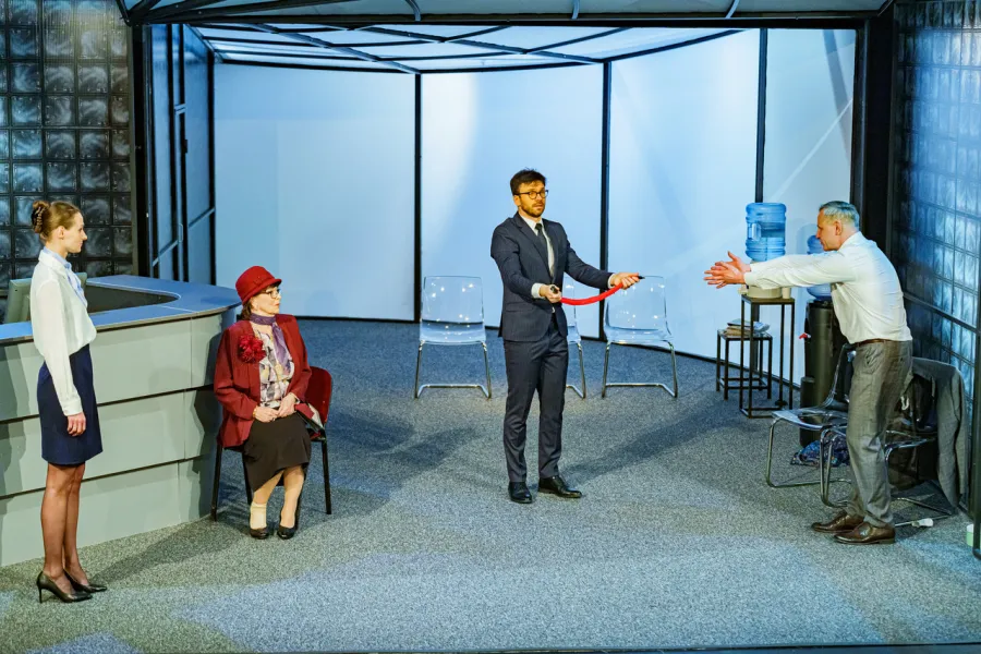 Na scenie teatralnej stoją cztery postacie: kobieta po lewej stronie, siedząca starsza kobieta w czerwonym kapeluszu, stojący w centrum mężczyzna trzymający czerwony pasek oraz mężczyzna w białej koszuli wyciągający ręce po prawej stronie. W tle widać minimalistyczną scenografię przypominającą nowoczesne biuro.