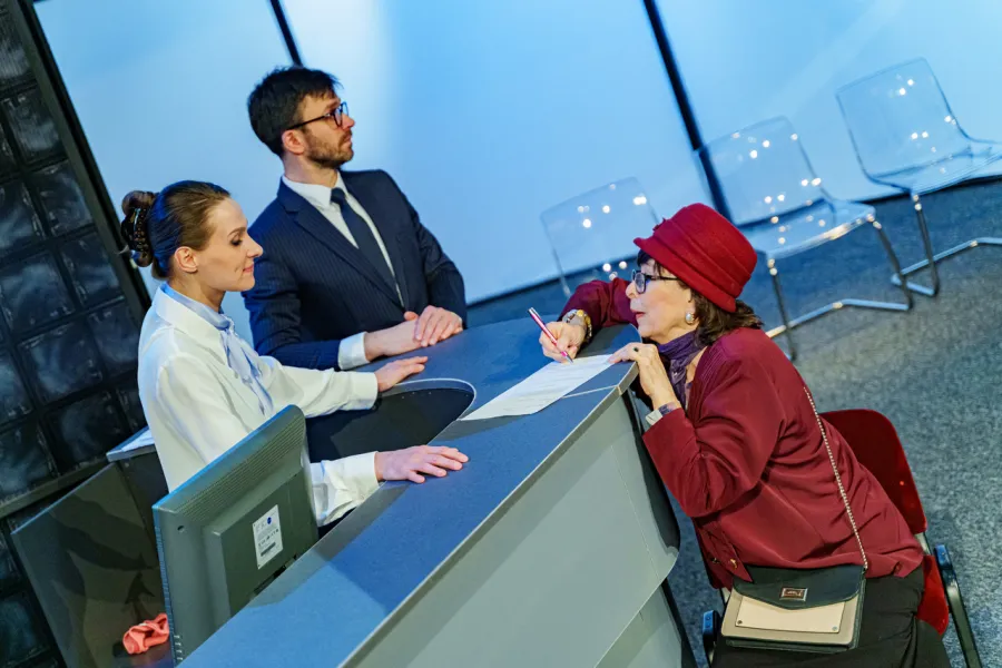 Na scenie teatralnej starsza kobieta w czerwonym kapeluszu siedzi przy recepcji i podpisuje dokumenty. Obok lady stoją kobieta w białej koszuli oraz mężczyzna w garniturze i okularach.