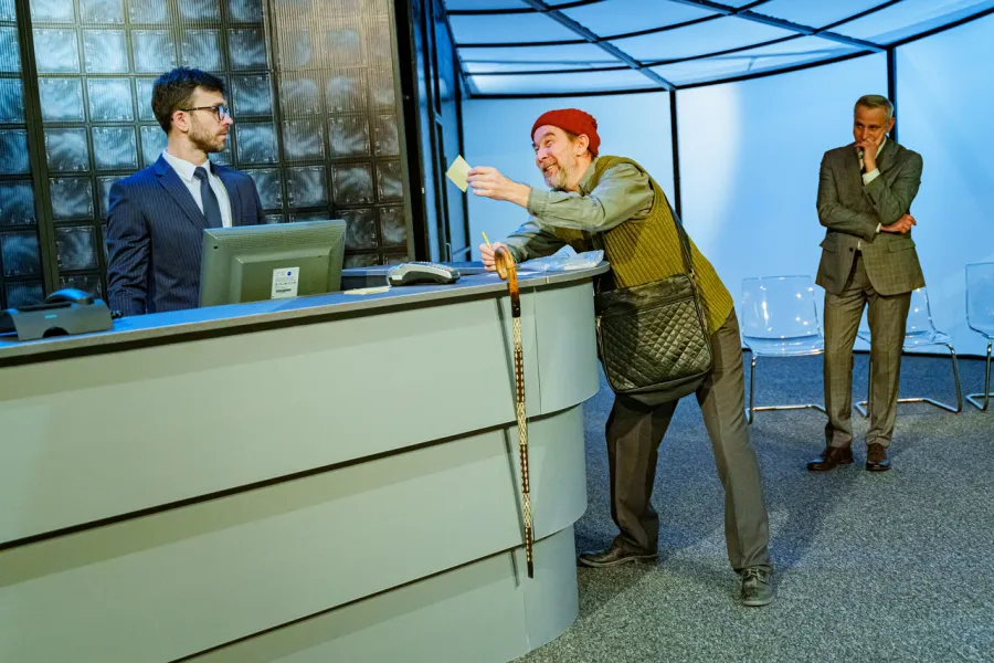 Na scenie teatralnej mężczyzna w czerwonej czapce i z torbą przewieszoną przez ramię podaje coś pracownikowi siedzącemu za ladą recepcji. W tle z boku stoi inny mężczyzna w garniturze, przyglądając się tej scenie z ręką na brodzie.