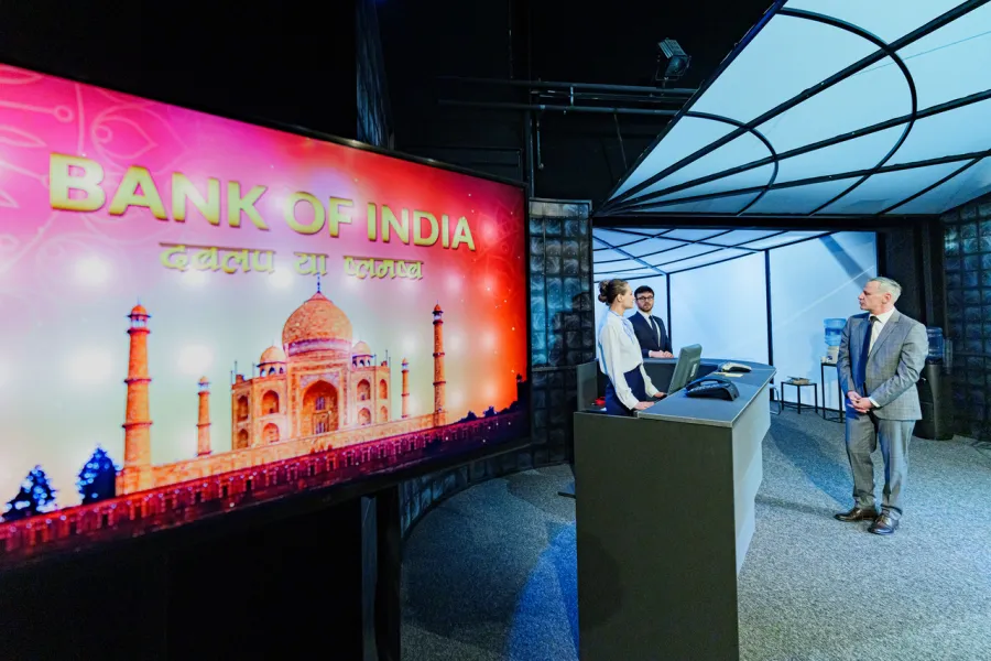 Na pierwszym planie widoczny jest duży ekran wyświetlający napis „BANK OF INDIA” oraz grafikę przedstawiającą Tadź Mahal. W tle, na scenie przypominającej nowoczesne biuro lub bank, stoją pracownicy za ladą oraz mężczyzna w garniturze.