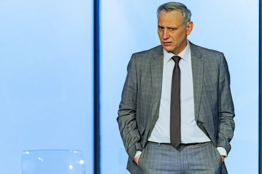 Na scenie teatralnej mężczyzna w szarym garniturze, białej koszuli i z ciemnym krawatem stoi z rękoma w kieszeniach spodni, patrząc przed siebie z powagą. W tle widoczna jest jasna, minimalistyczna ściana scenerii.