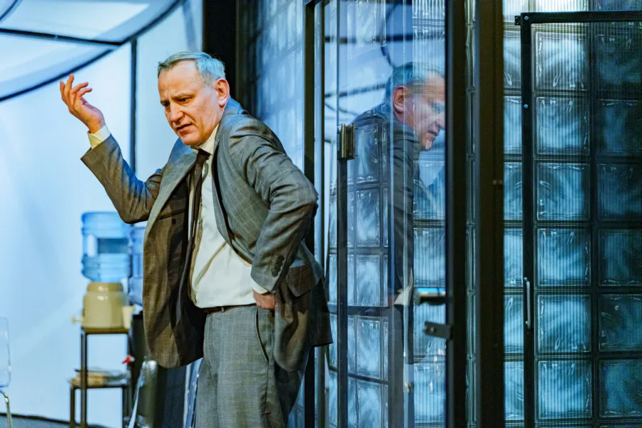 Na scenie teatralnej mężczyzna w szarym garniturze stoi w nowoczesnym wnętrzu, wykonując gest uniesioną ręką i patrząc w bok z przejęciem. W pobliżu odbija się w szklanej ścianie z luksferów.