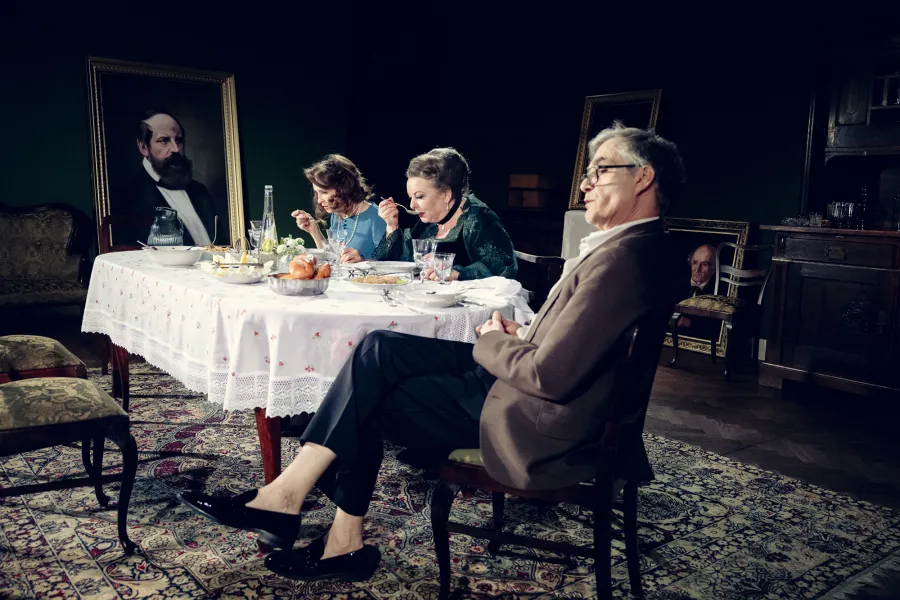 Scena teatralna przedstawia troje aktorów siedzących przy stole w elegancko urządzonej jadalni. Dwie kobiety jedzą posiłek, podczas gdy mężczyzna siedzący na pierwszym planie patrzy w bok. W tle widoczne są obrazy w złotych ramach oraz ciemny kredens.